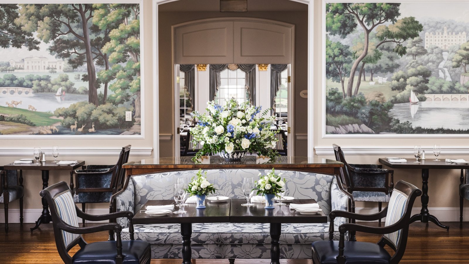 An elegant table is set for breakfast amidst a room decorated with scenic murals and sunlight from the large windows looking out to the gardens of the Williamsburg Inn.