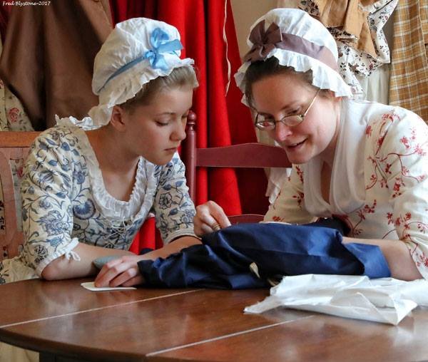 A photo of a girl and woman in 18th-century clothing looking at navy blue fabric at a round, wooden table