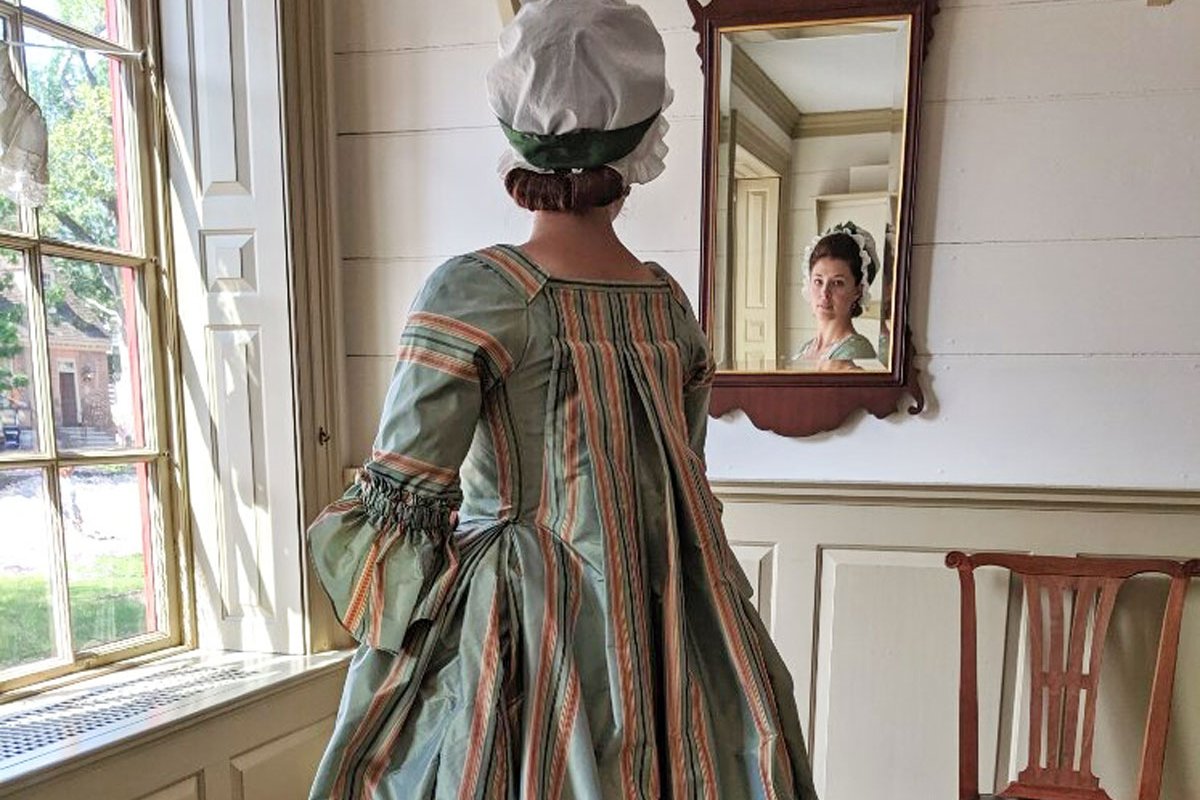 A photo of a woman dressed in an 18th-century blue and pink gown, looking at herself in the mirror