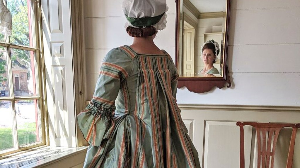 A photo of a woman dressed in an 18th-century blue and pink gown, looking at herself in the mirror