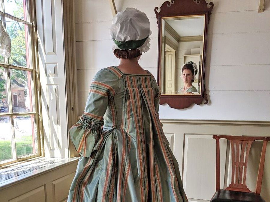 A photo of a woman dressed in an 18th-century blue and pink gown, looking at herself in the mirror