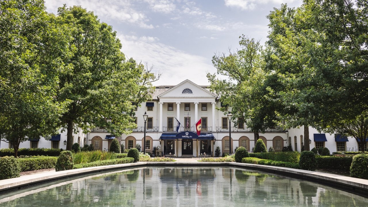 The front view of The Williamsburg Inn. As you approach this iconic hotel, you'll be greeted by lines of trees, elegant landmark signage, a reflection pond, and a royal blue awning supported by brass poles.