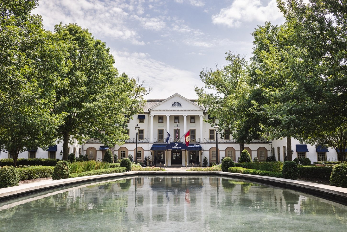 The front view of The Williamsburg Inn. As you approach this iconic hotel, you'll be greeted by lines of trees, elegant landmark signage, a reflection pond, and a royal blue awning supported by brass poles.