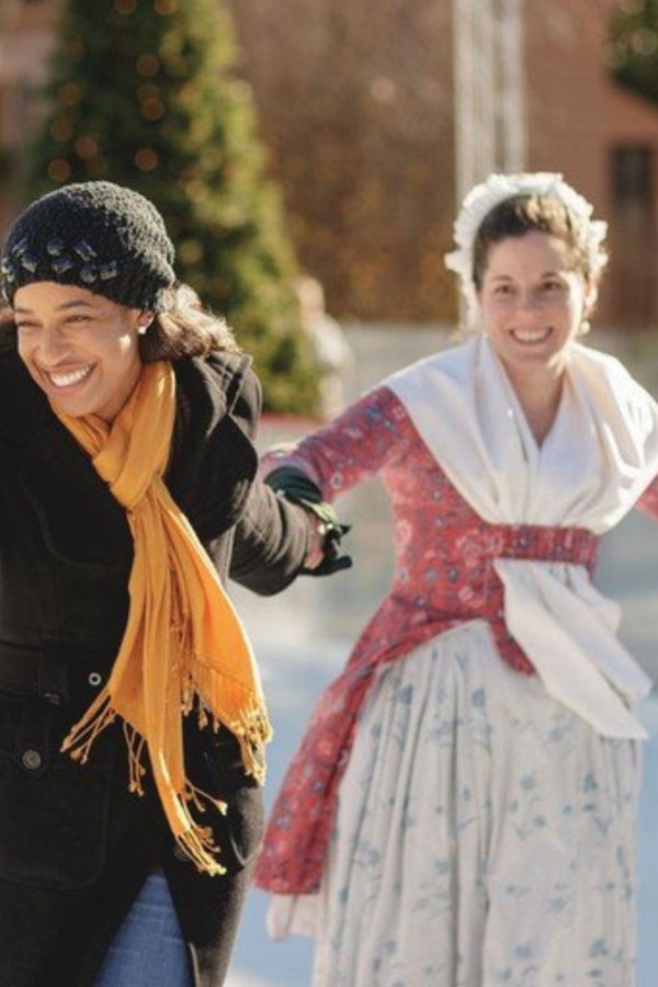 A woman and a costumed interpreter hold hands as they skate on the ice rink