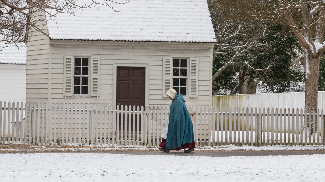 A person in a teal cloak walks past a white fence on a snowy day