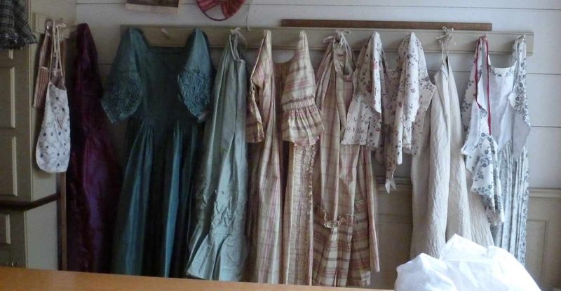 A photo of a row of 18th century dresses and other garments on wall hooks.