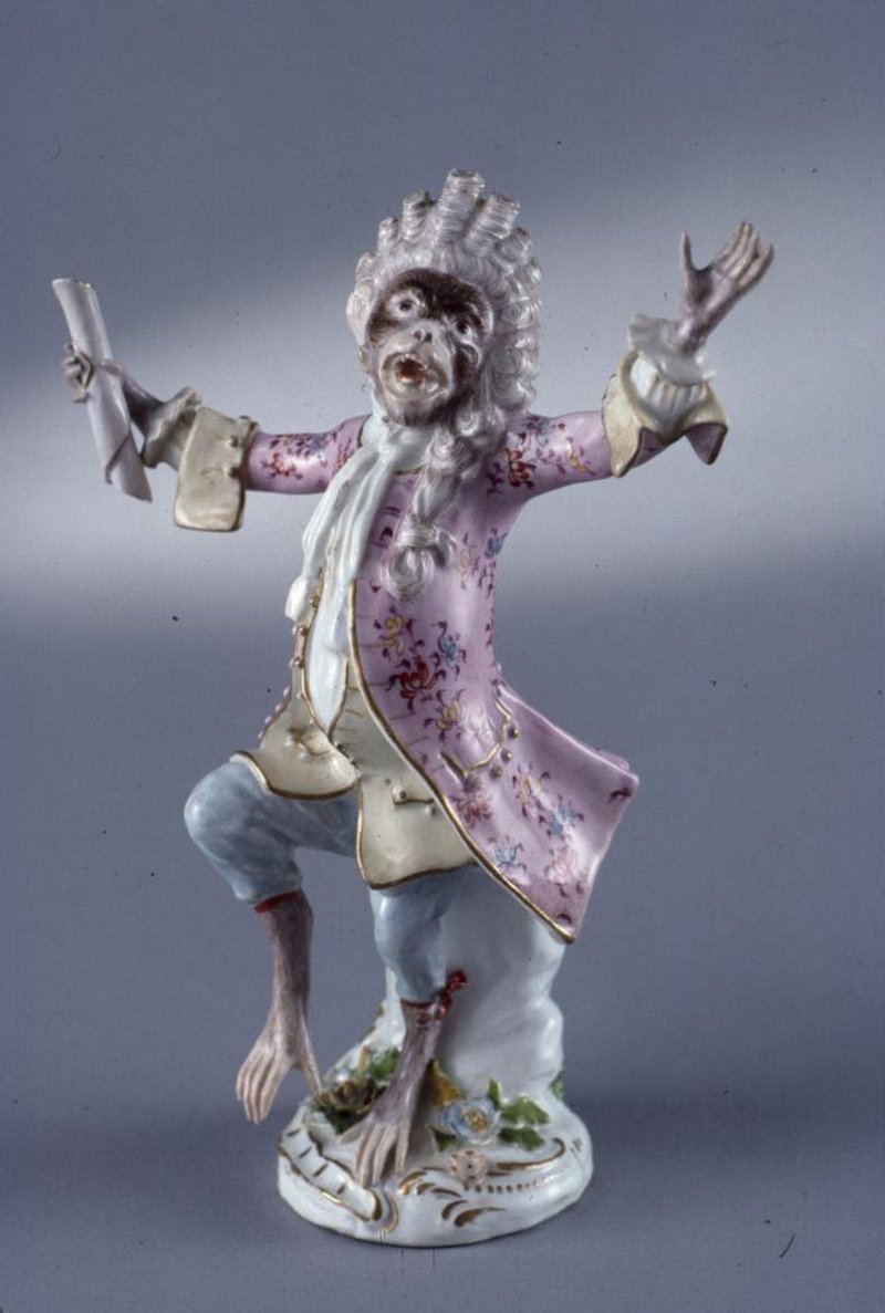 A photo of a porcelain monkey figure dressed in 18th century clothing, holding a scroll of paper.